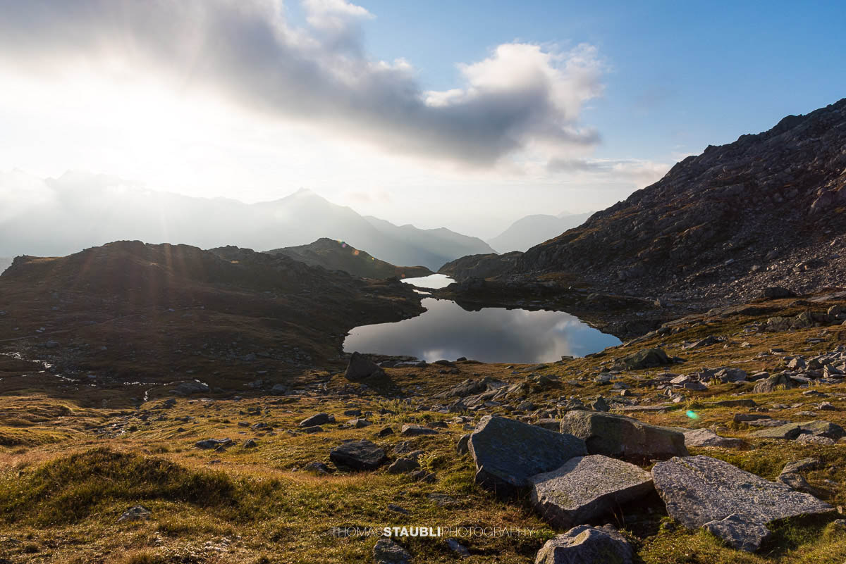 Sonnenaufgang an den Laghi della Valletta: ruhiger Bergsee mit Spiegelung von Himmel und Wolken, sanftes Morgenlicht über einer kargen Hochalpenlandschaft im Tessin.