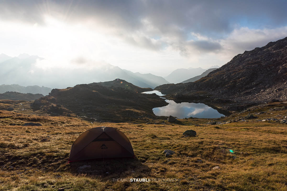 Sonnenaufgang an den Laghi della Valletta: ruhiger Bergsee mit Spiegelung von Himmel und Wolken, sanftes Morgenlicht über einer kargen Hochalpenlandschaft im Tessin.