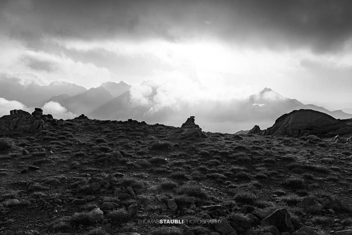 Schwarzweissaufnahme der Laghi della Valletta: ruhiger Bergsee mit Spiegelung dunkler Wolken, felsige Ufer und Silhouetten ferner Gipfel in den Tessiner Alpen.