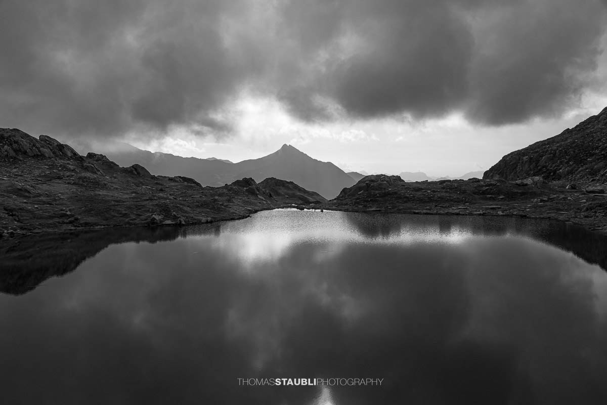 Schwarzweissaufnahme der Laghi della Valletta: ruhiger Bergsee mit Spiegelung dunkler Wolken, felsige Ufer und Silhouetten ferner Gipfel in den Tessiner Alpen.