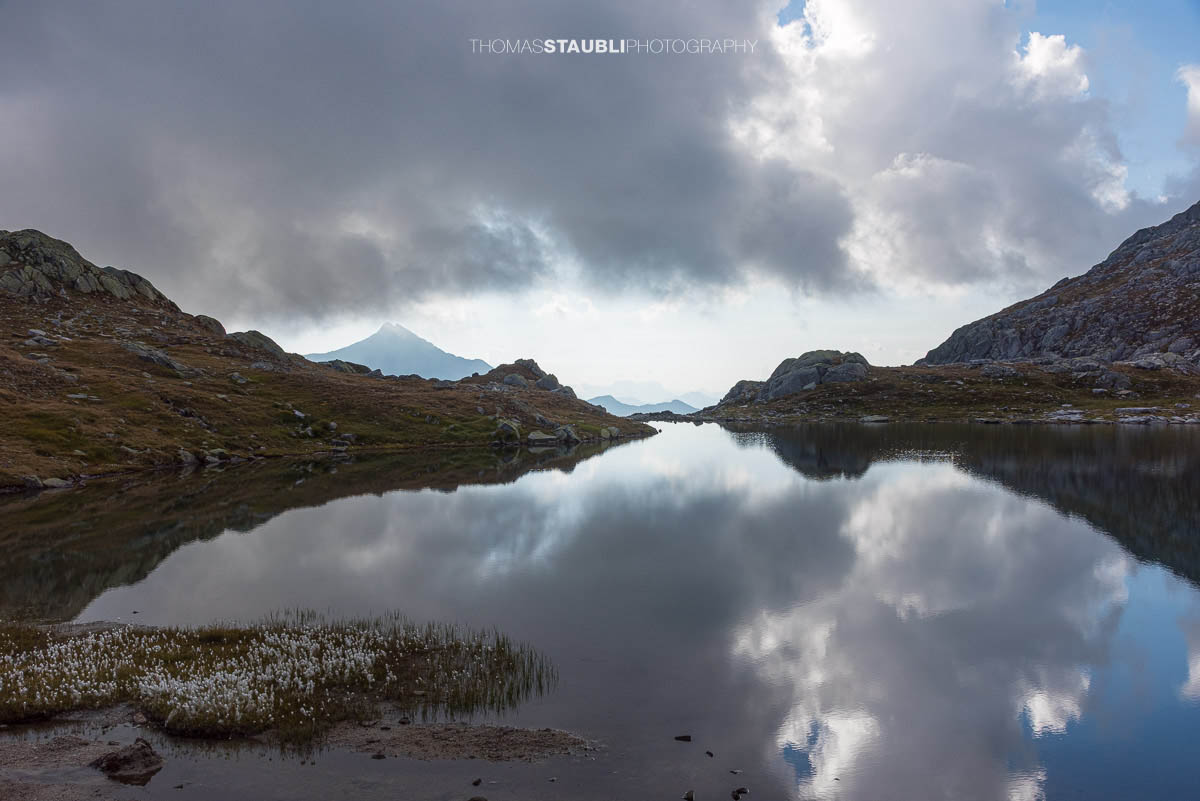Ruhiger Laghi della Valletta mit Spiegelung dunkler Wolken, felsige Ufer und Silhouetten ferner Gipfel in den Tessiner Alpen.