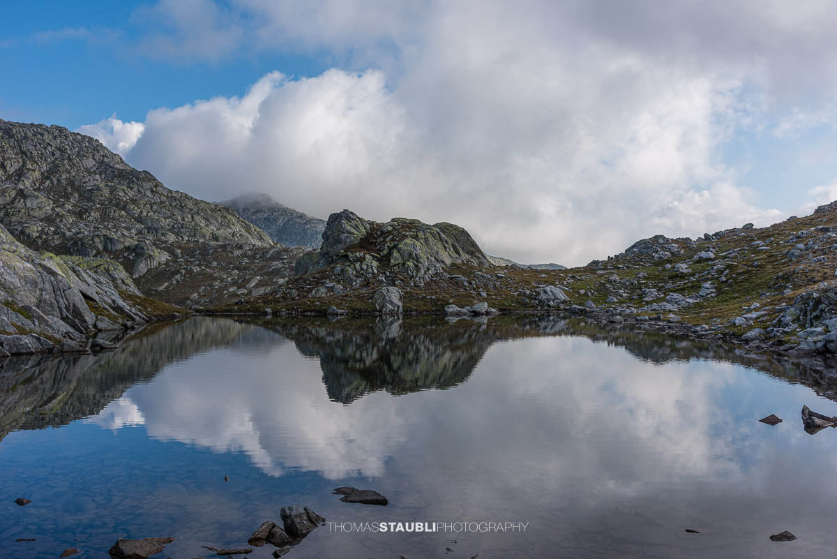 Ruhiger Laghi della Valletta mit Spiegelung dunkler Wolken, felsige Ufer und Silhouetten ferner Gipfel in den Tessiner Alpen.