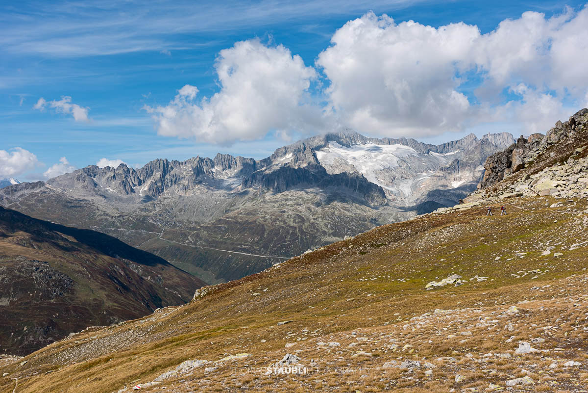 Blick vom Passo d'Orsirora über eine weite Hochalpenlandschaft zur Serpentine der Furkapassstrasse; im Hintergrund vergletscherte Gipfel und schroffe Grate unter blauem Himmel.