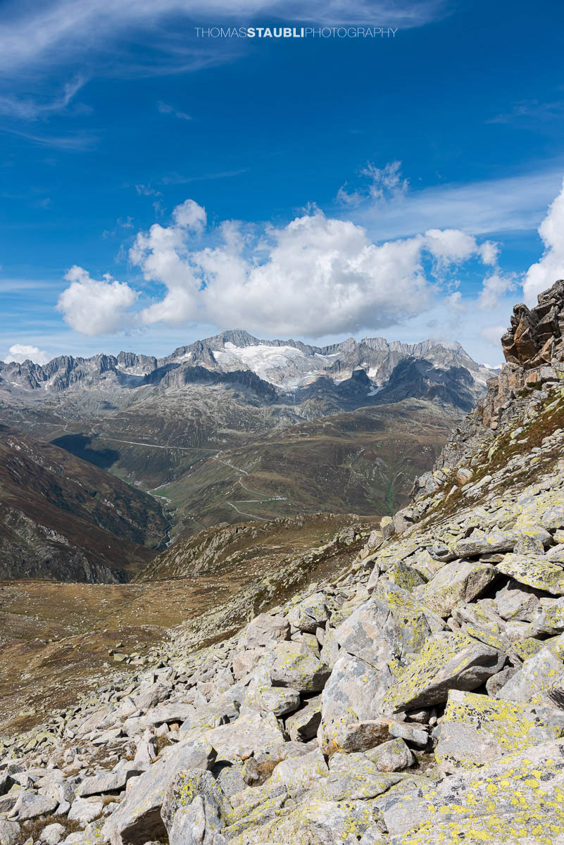 Blick vom Passo d'Orsirora über eine weite Hochalpenlandschaft zur Serpentine der Furkapassstrasse; im Hintergrund vergletscherte Gipfel und schroffe Grate unter blauem Himmel.