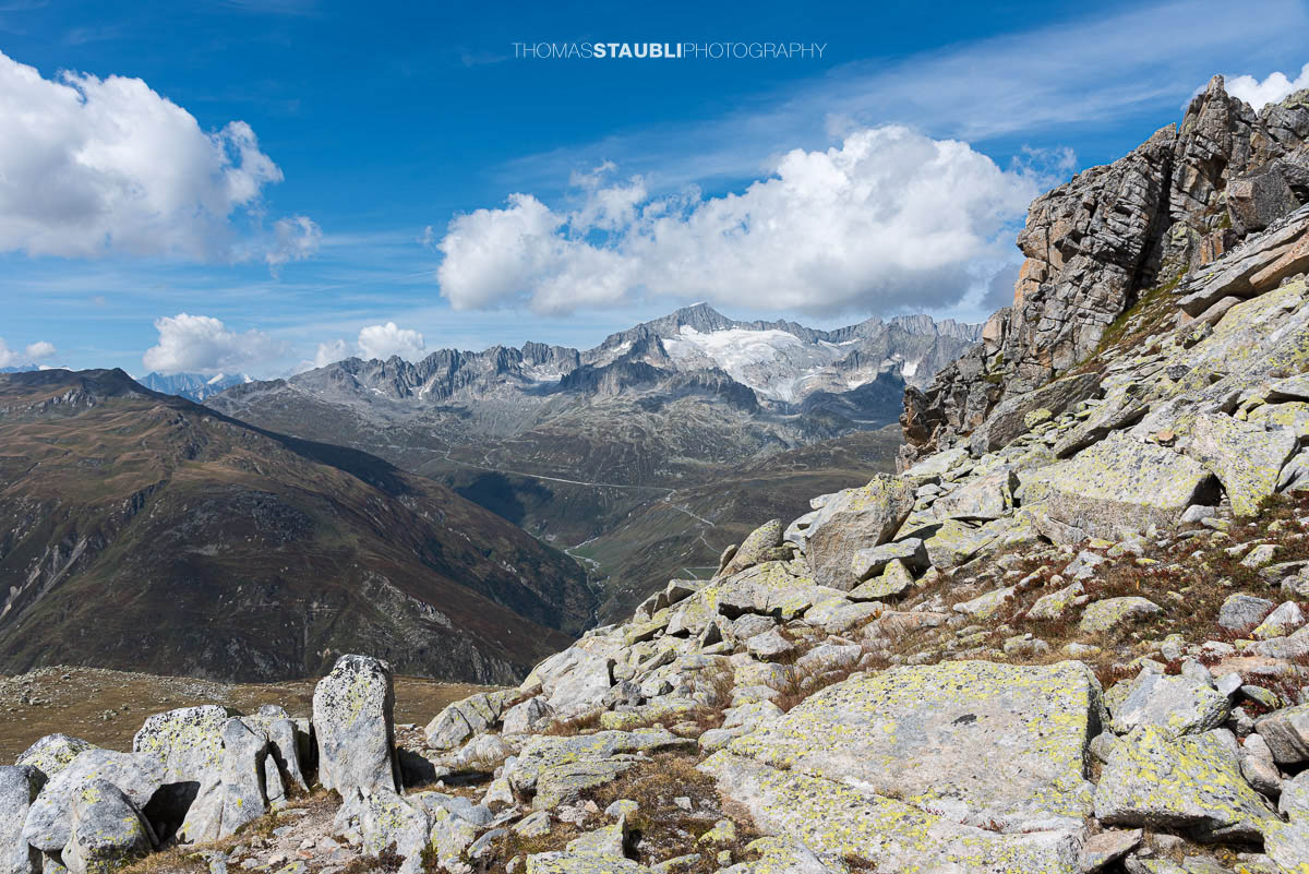 Blick vom Passo d'Orsirora über eine weite Hochalpenlandschaft zur Serpentine der Furkapassstrasse; im Hintergrund vergletscherte Gipfel und schroffe Grate unter blauem Himmel.