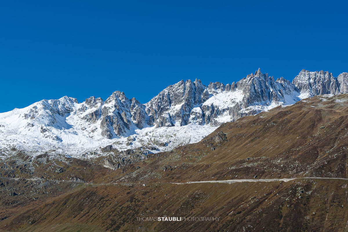 Blick zur Furkapassstrasse im Urserental mit Furkahorn und Sidelenhorn im Hintergrund, herbstliche Hänge und schneebedeckte Gipfel unter klarem Himmel.