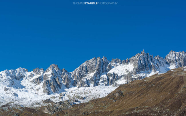 Blick zur Furkapassstrasse im Urserental mit Furkahorn und Sidelenhorn im Hintergrund, herbstliche Hänge und schneebedeckte Gipfel unter klarem Himmel.