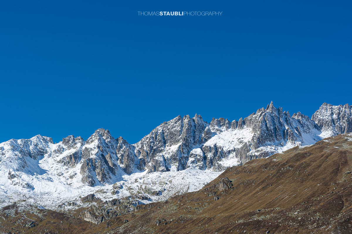 Blick zur Furkapassstrasse im Urserental mit Furkahorn und Sidelenhorn im Hintergrund, herbstliche Hänge und schneebedeckte Gipfel unter klarem Himmel.