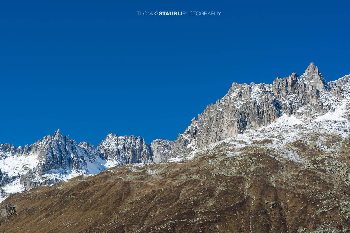 Blick zur Furkapassstrasse im Urserental mit Furkahorn und Sidelenhorn im Hintergrund, herbstliche Hänge und schneebedeckte Gipfel unter klarem Himmel.