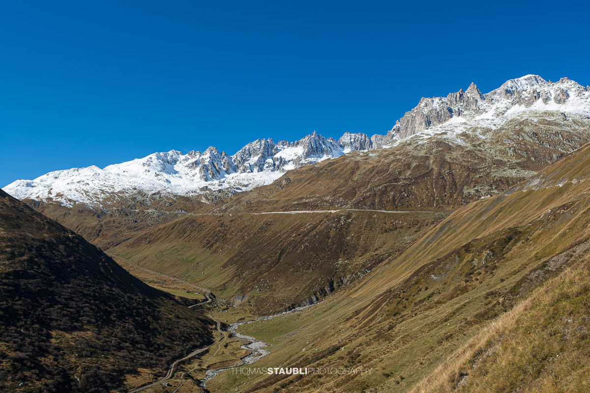 Blick zur Furkapassstrasse im Urserental mit Furkahorn und Galenstock im Hintergrund, herbstliche Hänge und schneebedeckte Gipfel unter klarem Himmel.