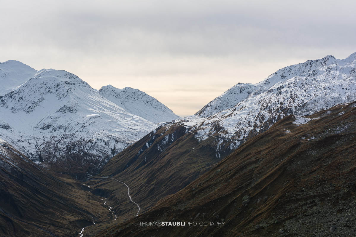 Blick über das Urserental zum Furkapass mit dem Hotel Furkablick auf der Passhöhe, umgeben von schneebedeckten Bergen und herbstlichen Hängen.