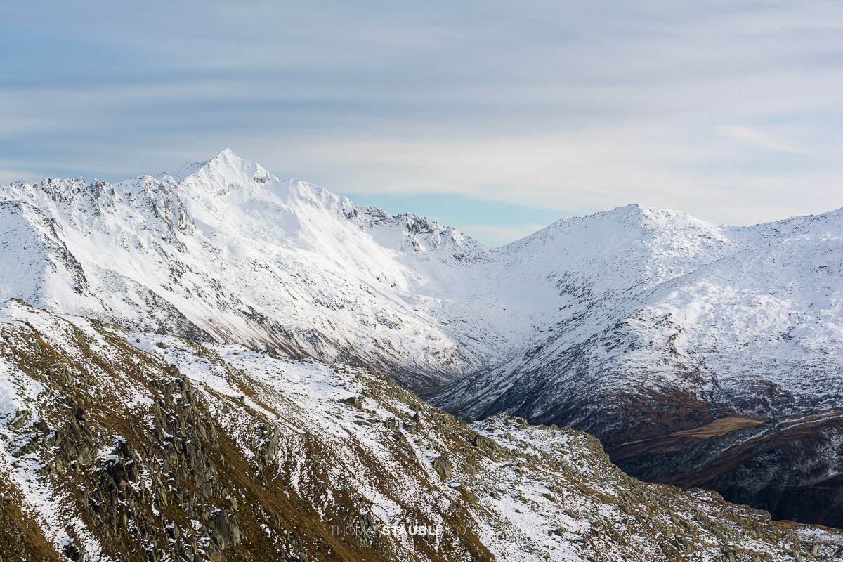 Blick zum Pizzo Lucendro im Urserental, verschneite Gipfel und herbstlich gefärbte Hänge im Übergang zum Winter.