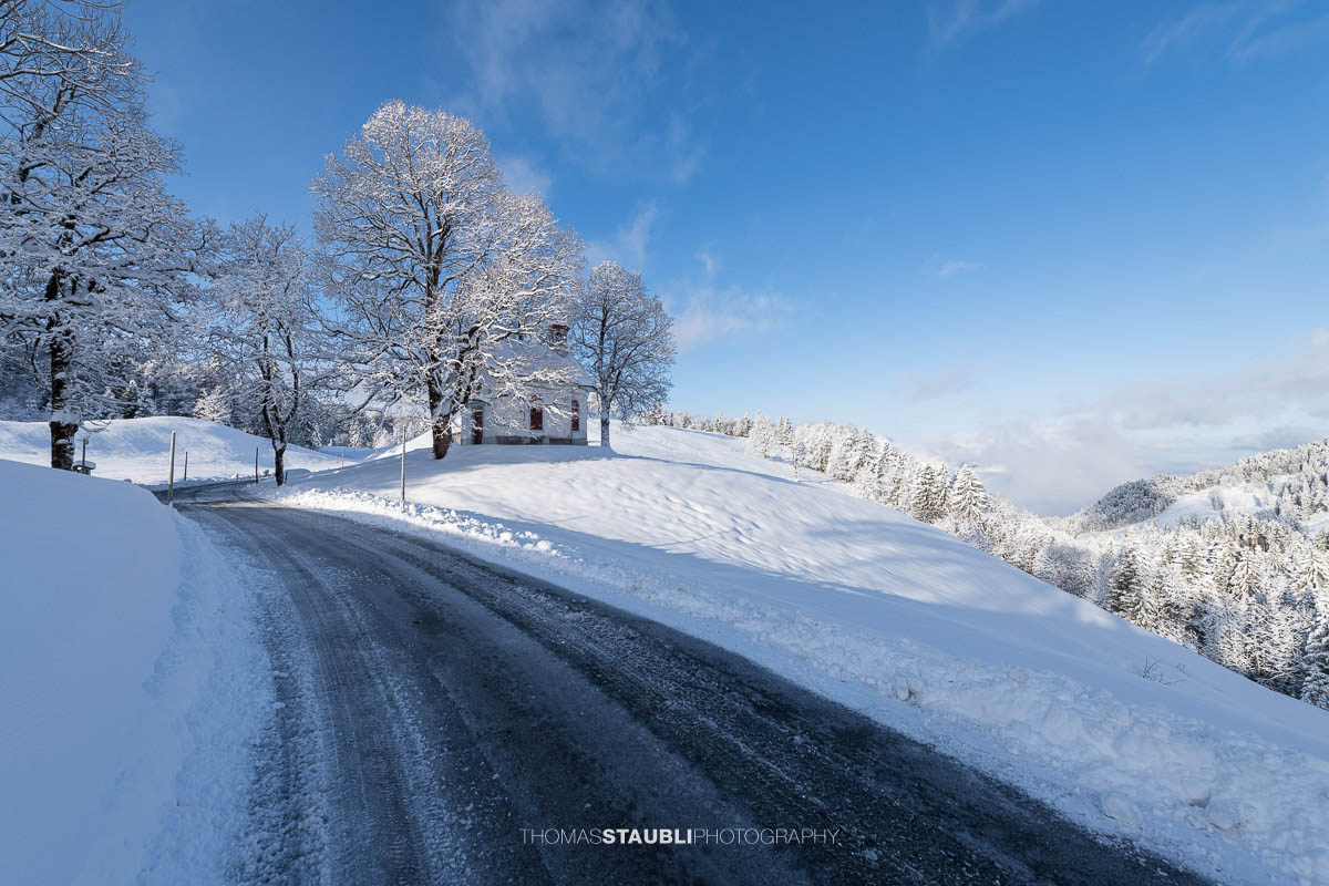 Kleine Kapelle St. Anna im Gründel, umgeben von schneebedeckten Wiesen und alten Bäumen, mit roter Eingangstür in ruhiger Winterlandschaft.
