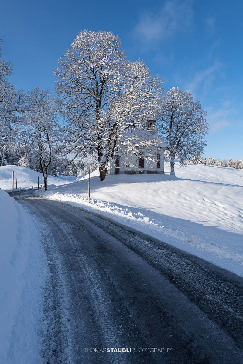 Kleine Kapelle St. Anna im Gründel, umgeben von schneebedeckten Wiesen und alten Bäumen, mit roter Eingangstür in ruhiger Winterlandschaft.