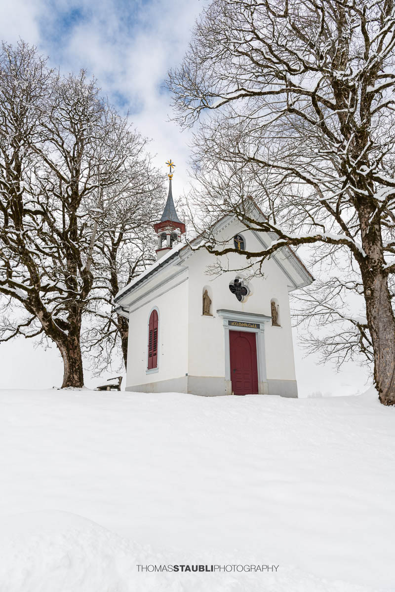 Kleine Kapelle St. Anna im Gründel, umgeben von schneebedeckten Wiesen und alten Bäumen, mit roter Eingangstür in ruhiger Winterlandschaft.