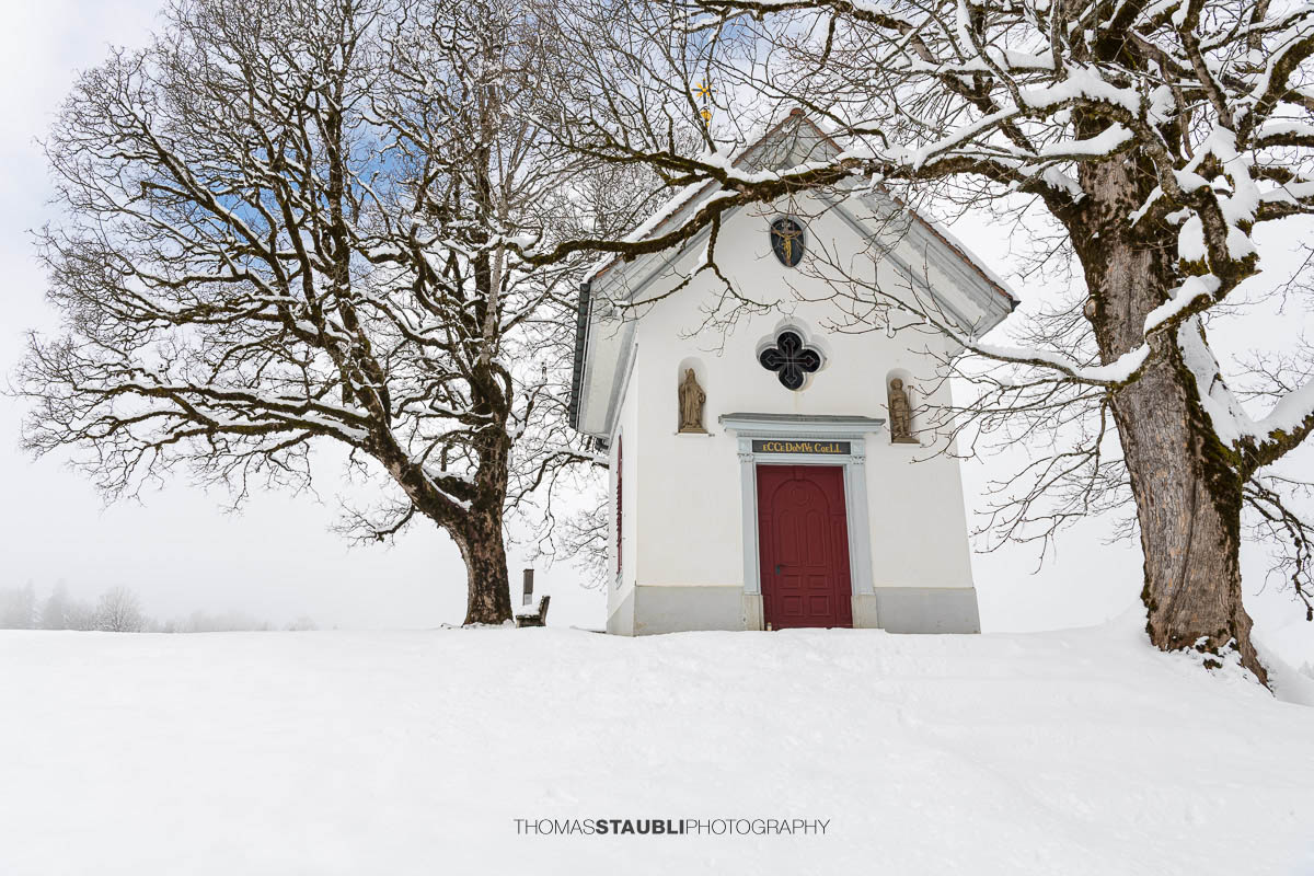 Kleine Kapelle St. Anna im Gründel, umgeben von schneebedeckten Wiesen und alten Bäumen, mit roter Eingangstür in ruhiger Winterlandschaft.