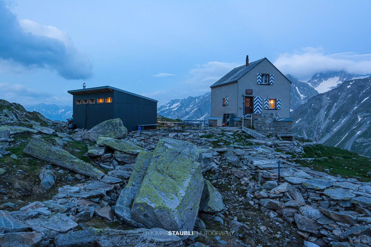 Dammahütte in den Urner Alpen bei Abenddämmerung, beleuchtete Fenster inmitten felsiger Berglandschaft unter wolkigem Himmel.