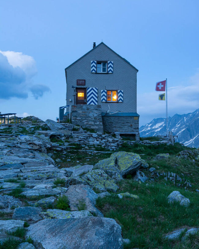Dammahütte in den Urner Alpen bei Abenddämmerung, beleuchtete Fenster inmitten felsiger Berglandschaft unter wolkigem Himmel.