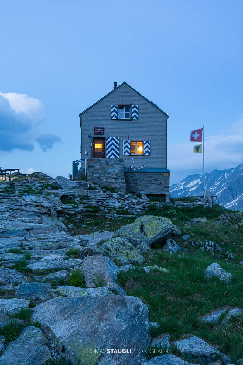 Dammahütte in den Urner Alpen bei Abenddämmerung, beleuchtete Fenster inmitten felsiger Berglandschaft unter wolkigem Himmel.