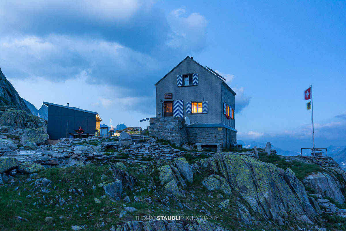 Dammahütte in den Urner Alpen bei Abenddämmerung, beleuchtete Fenster inmitten felsiger Berglandschaft unter wolkigem Himmel.