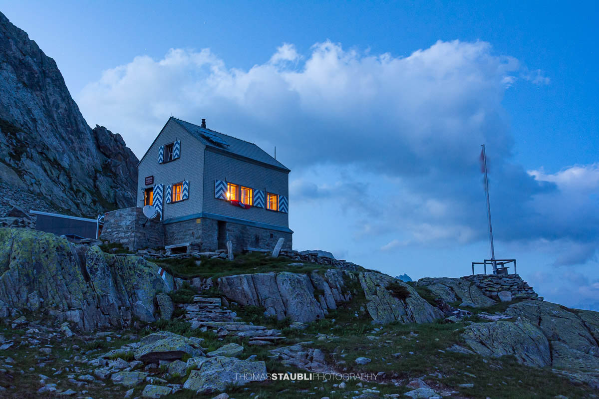 Dammahütte in den Urner Alpen bei Abenddämmerung, beleuchtete Fenster inmitten felsiger Berglandschaft unter wolkigem Himmel.