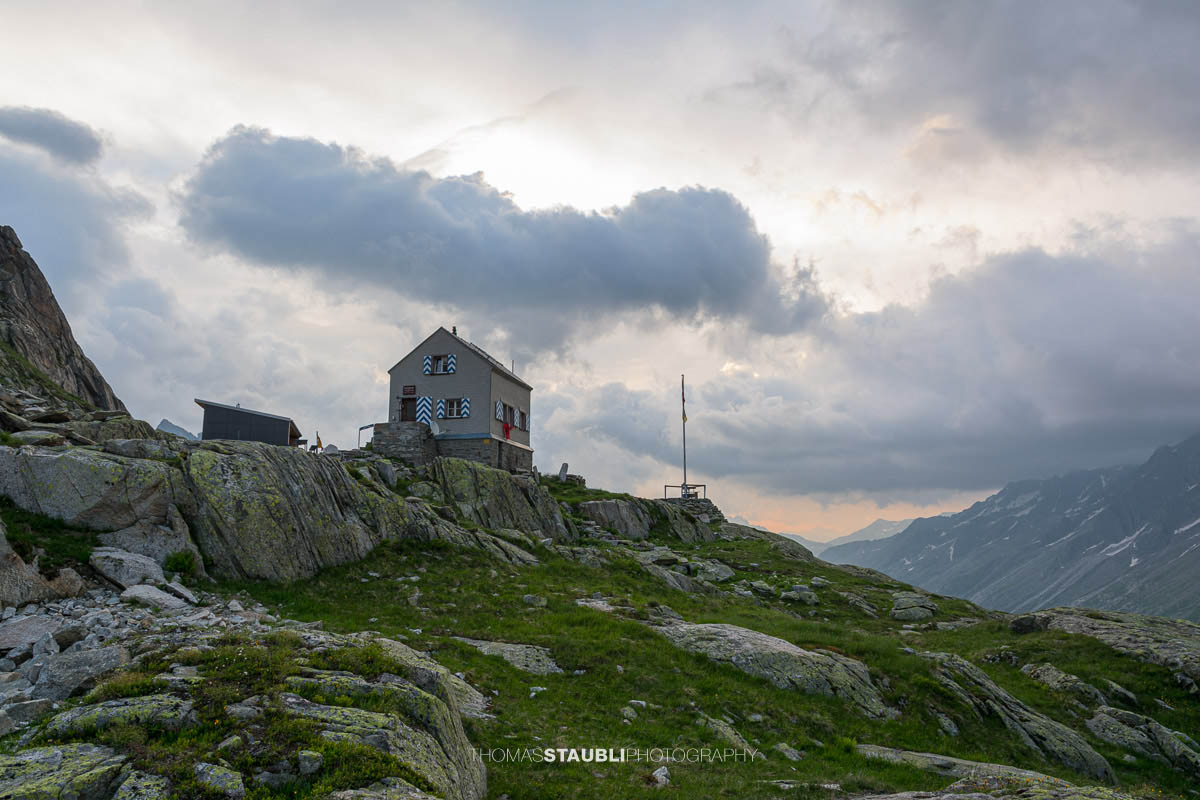 Dammahütte in den Urner Alpen im Kanton Uri bei Morgendämmerung, Hütte auf Felsen mit wehender Fahne unter wolkigem Himmel im ersten Licht.