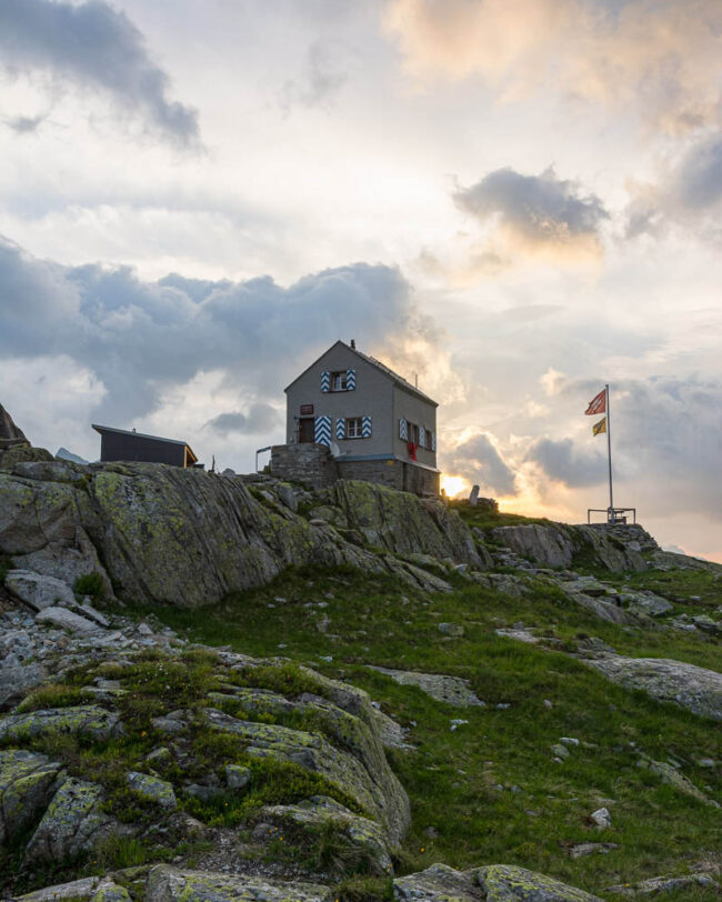 Dammahütte in den Urner Alpen im Kanton Uri bei Morgendämmerung, Hütte auf Felsen mit wehender Fahne unter wolkigem Himmel im ersten Licht.