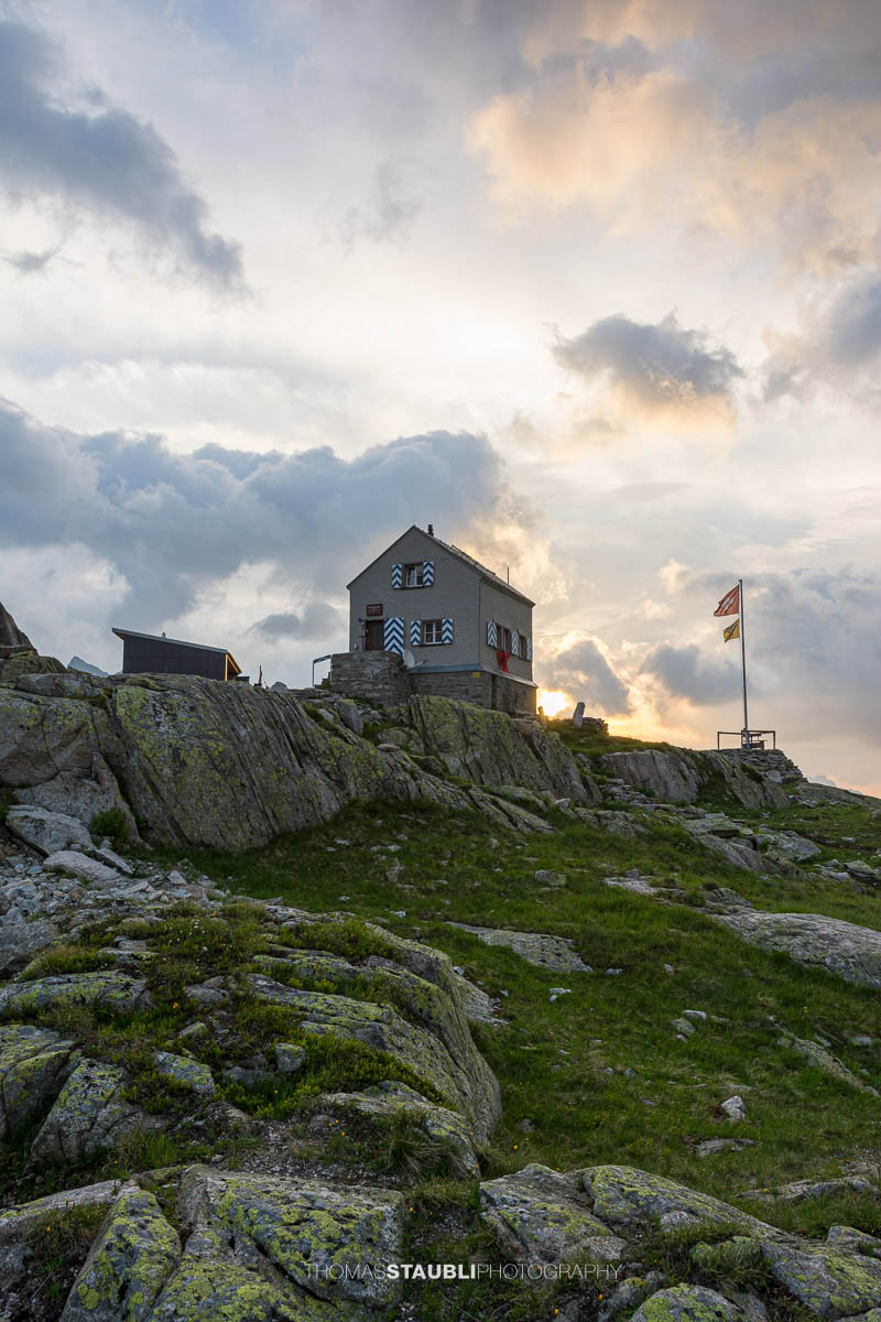 Dammahütte in den Urner Alpen im Kanton Uri bei Morgendämmerung, Hütte auf Felsen mit wehender Fahne unter wolkigem Himmel im ersten Licht.