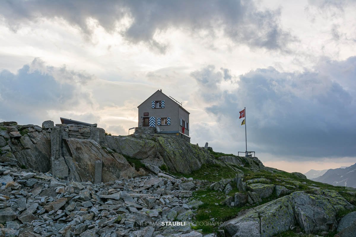 Dammahütte in den Urner Alpen im Kanton Uri bei Morgendämmerung, Hütte auf Felsen mit wehender Fahne unter wolkigem Himmel im ersten Licht.