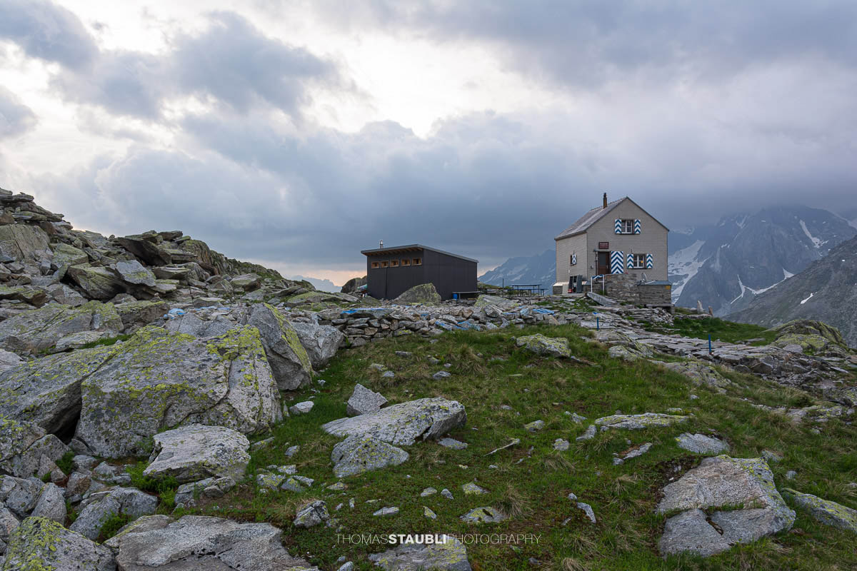 Dammahütte in den Urner Alpen im Kanton Uri bei Morgendämmerung, Hütte auf Felsen mit wehender Fahne unter wolkigem Himmel im ersten Licht.