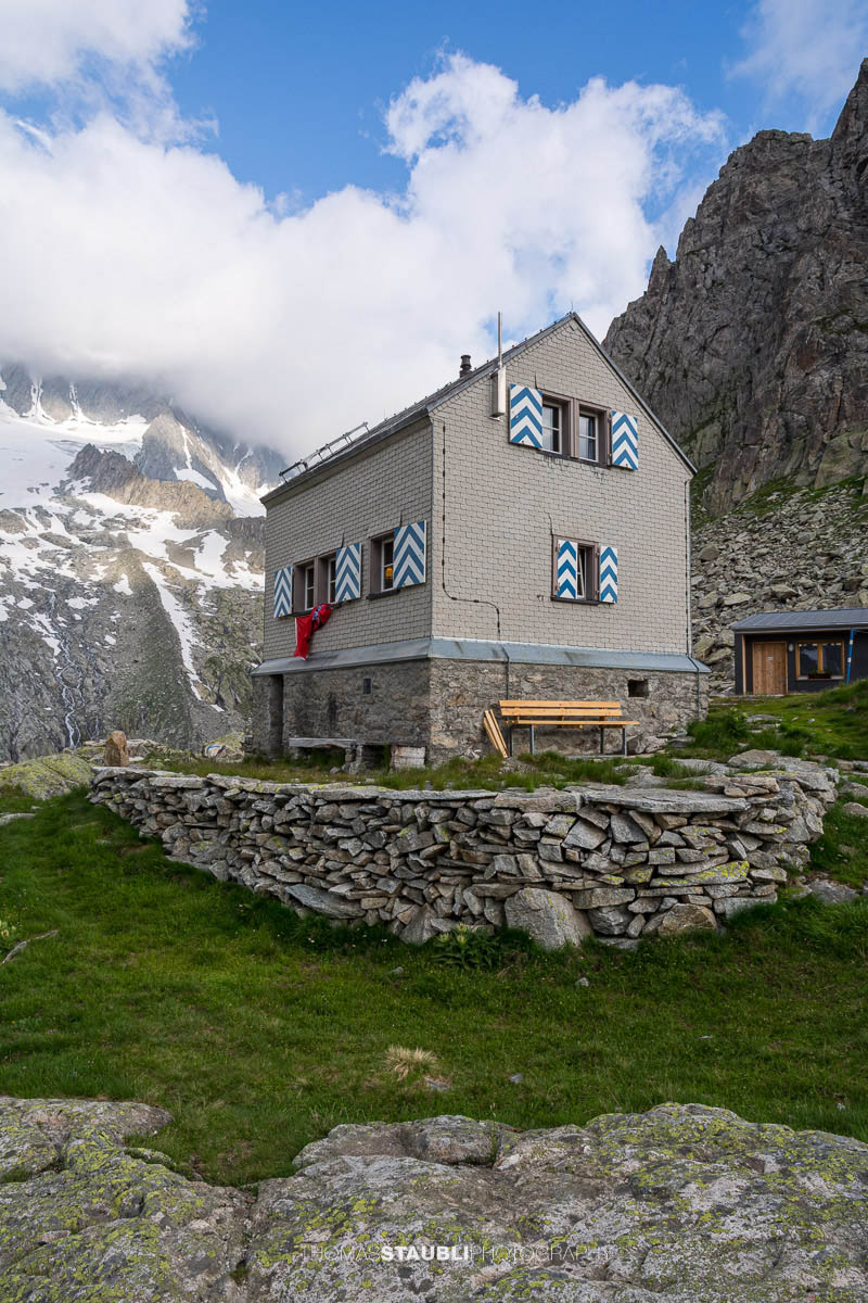 Dammahütte in den Urner Alpen im Kanton Uri bei Morgendämmerung, Hütte auf Felsen mit wehender Fahne unter wolkigem Himmel im ersten Licht.