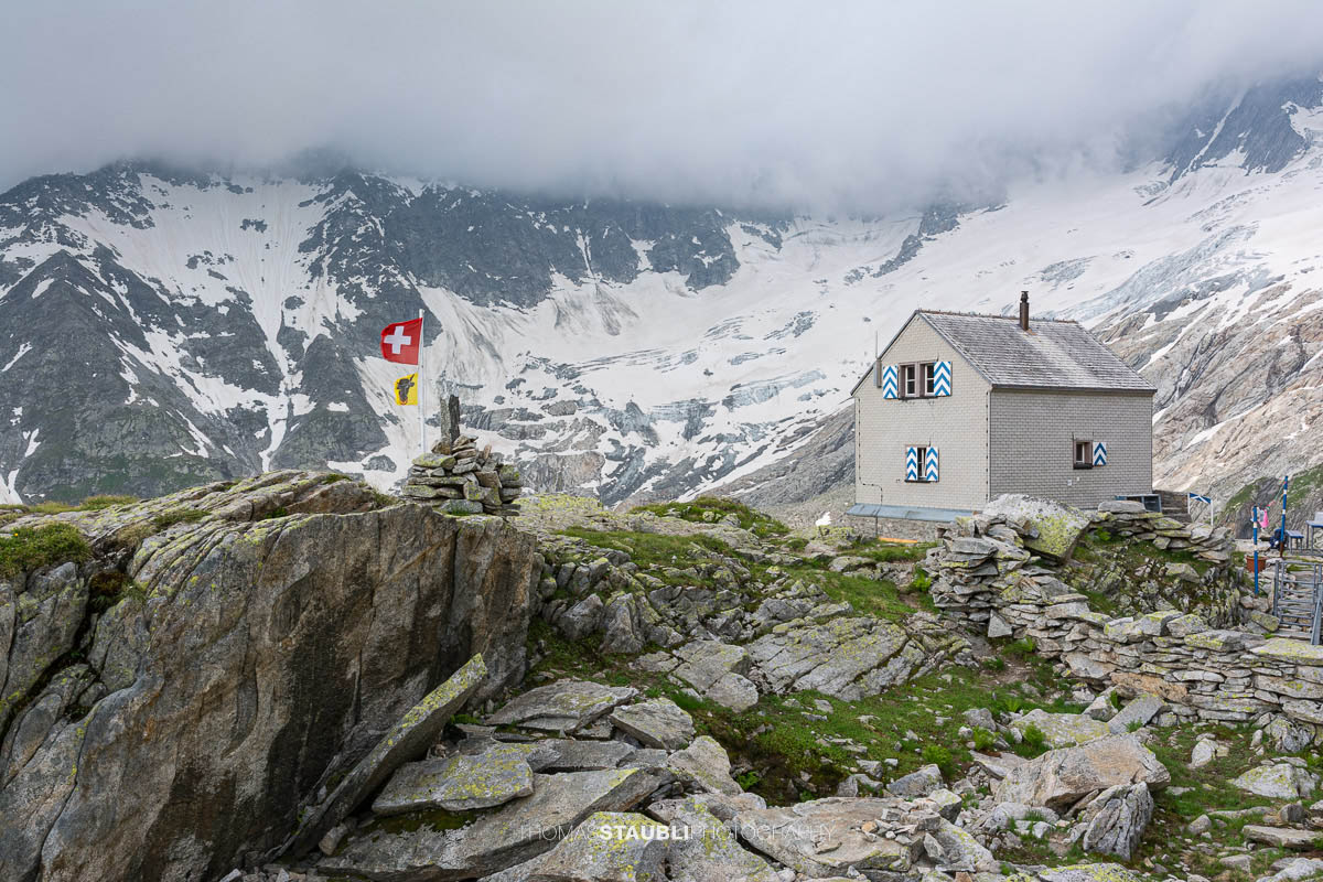 Dammahütte in den Urner Alpen im Kanton Uri bei Morgendämmerung, Hütte auf Felsen mit wehender Fahne unter wolkigem Himmel im ersten Licht.
