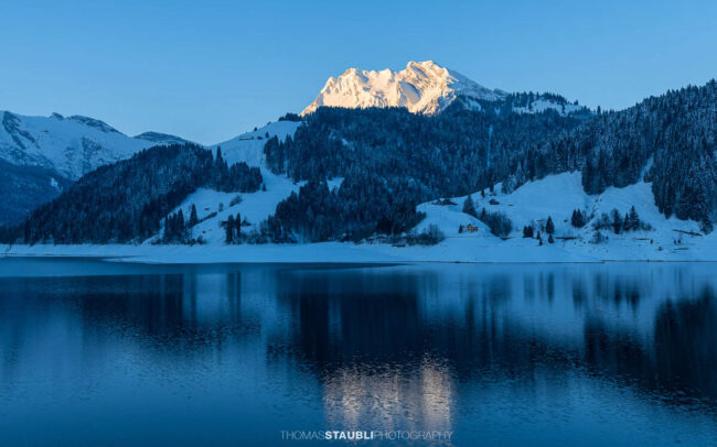 Morgenstimmung am Wägitalersee im Kanton Schwyz mit spiegelnder Wasseroberfläche und dem Fluebrig im ersten Sonnenlicht im Hintergrund.
