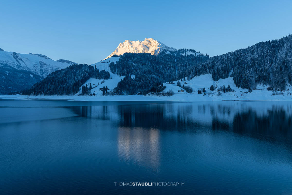 Morgenstimmung am Wägitalersee im Kanton Schwyz mit spiegelnder Wasseroberfläche und dem Fluebrig im ersten Sonnenlicht im Hintergrund.