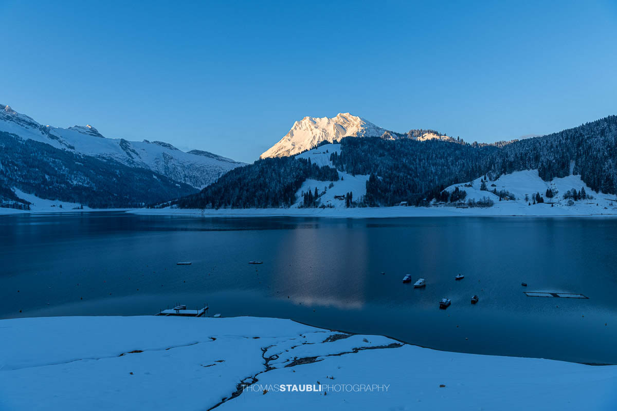 Morgenstimmung am Wägitalersee im Kanton Schwyz mit spiegelnder Wasseroberfläche und dem Fluebrig im ersten Sonnenlicht im Hintergrund.