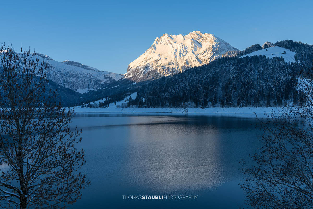 Morgenstimmung am Wägitalersee im Kanton Schwyz mit spiegelnder Wasseroberfläche und dem Fluebrig im ersten Sonnenlicht im Hintergrund.