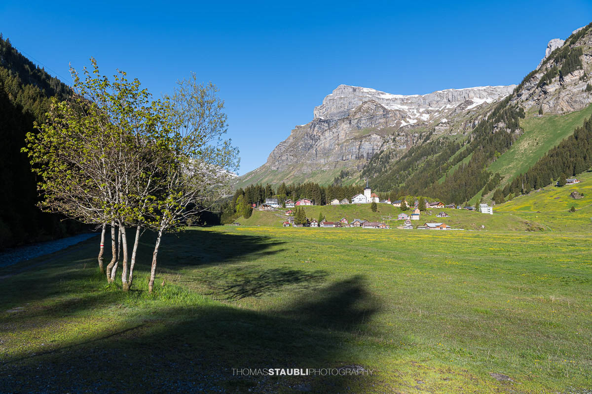 Blick über den Urnerboden im Kanton Uri mit der Kirche St. Erhard, grünen Wiesen und den markanten Bergflanken der Glarner Alpen unter tiefblauem Himmel.