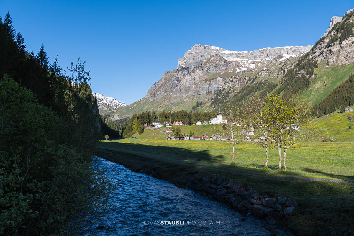 Wanderweg entlang dem Fätschbach auf dem Urnerboden im Kanton Uri mit der Kirche St. Erhard, grünen Wiesen und den markanten Bergflanken der Glarner Alpen unter tiefblauem Himmel.