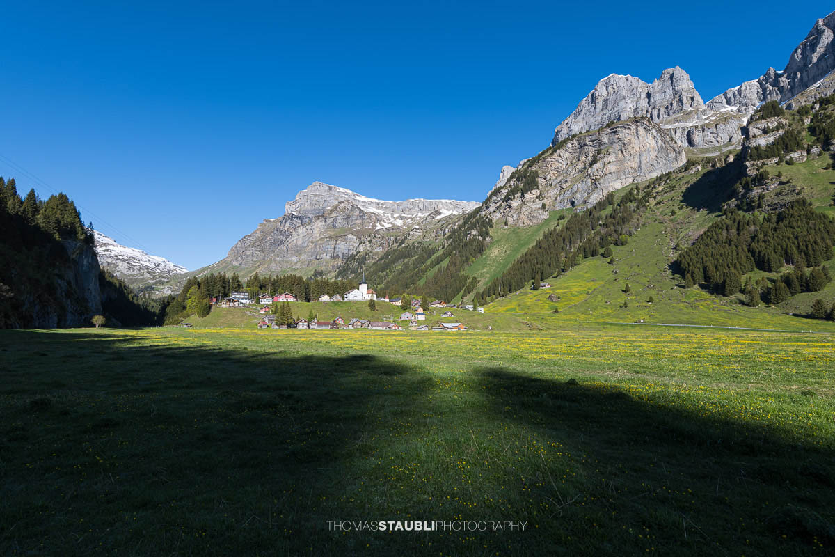 Blick über den Urnerboden im Kanton Uri mit der Kirche St. Erhard, grünen Wiesen und den markanten Bergflanken der Glarner Alpen unter tiefblauem Himmel.