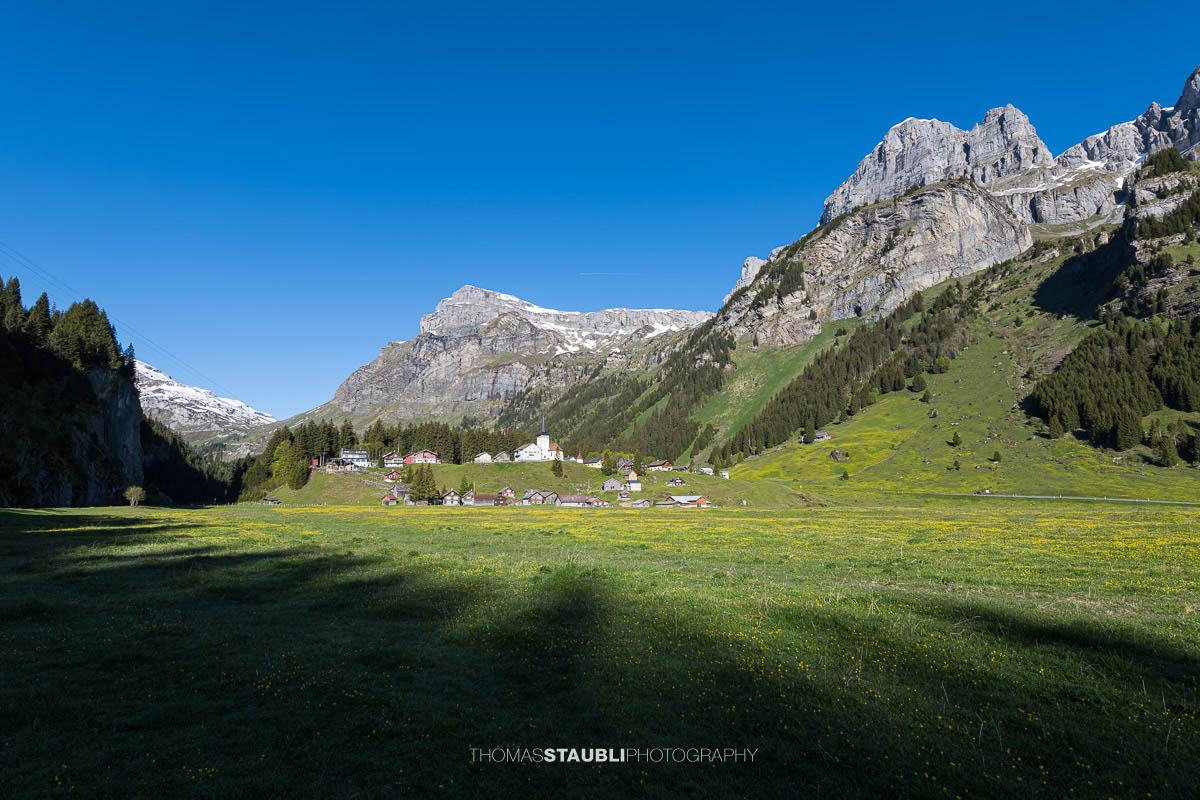 Blick über den Urnerboden im Kanton Uri mit der Kirche St. Erhard, grünen Wiesen und den markanten Bergflanken der Glarner Alpen unter tiefblauem Himmel.