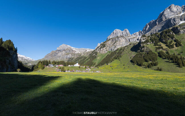 Blick über den Urnerboden im Kanton Uri mit der Kirche St. Erhard, grünen Wiesen und den markanten Bergflanken der Glarner Alpen unter tiefblauem Himmel.