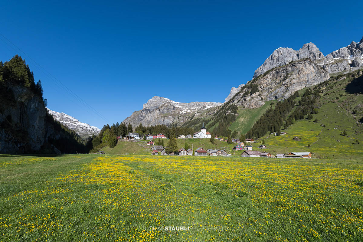 Blick über den Urnerboden im Kanton Uri mit der Kirche St. Erhard, grünen Wiesen und den markanten Bergflanken der Glarner Alpen unter tiefblauem Himmel.