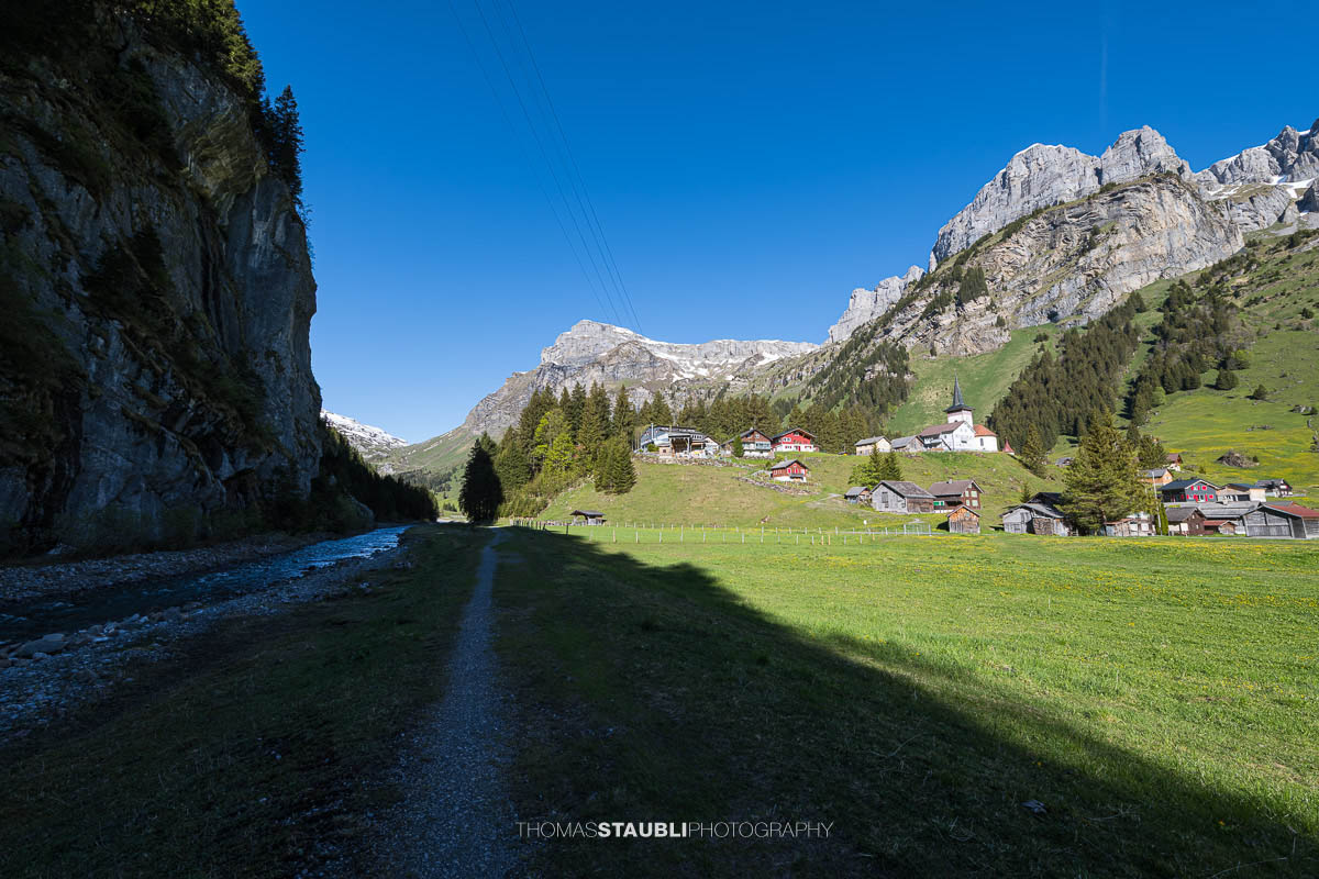 Wanderweg entlang dem Fätschbach auf dem Urnerboden im Kanton Uri mit der Kirche St. Erhard, grünen Wiesen und den markanten Bergflanken der Glarner Alpen unter tiefblauem Himmel.