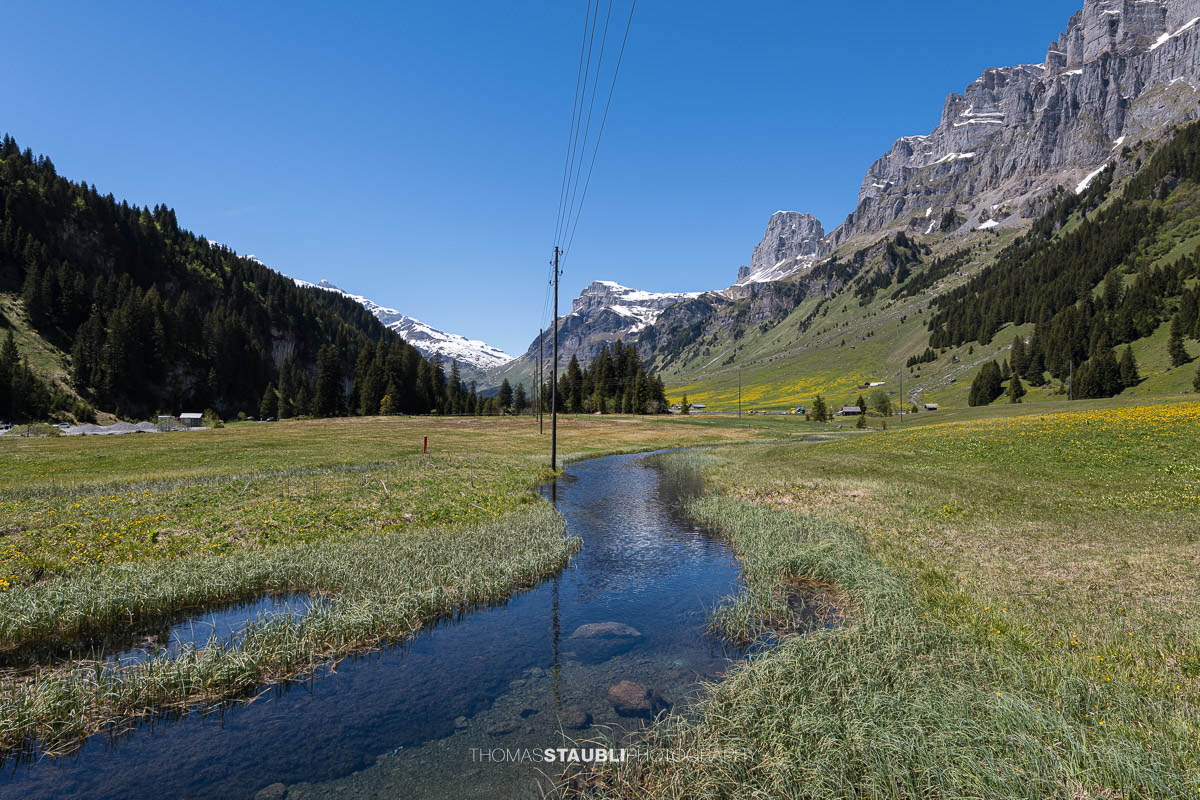 Kleiner Moorbach auf dem Urnerboden im Kanton Uri mit weiten Wiesen, steilen Felsflanken und den Bergen der Glarner Alpen unter klarem Himmel.