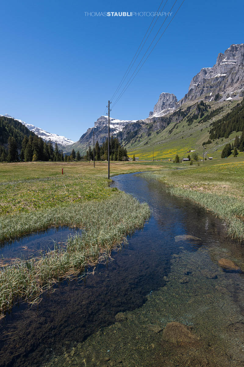 Kleiner Moorbach auf dem Urnerboden im Kanton Uri mit weiten Wiesen, steilen Felsflanken und den Bergen der Glarner Alpen unter klarem Himmel.