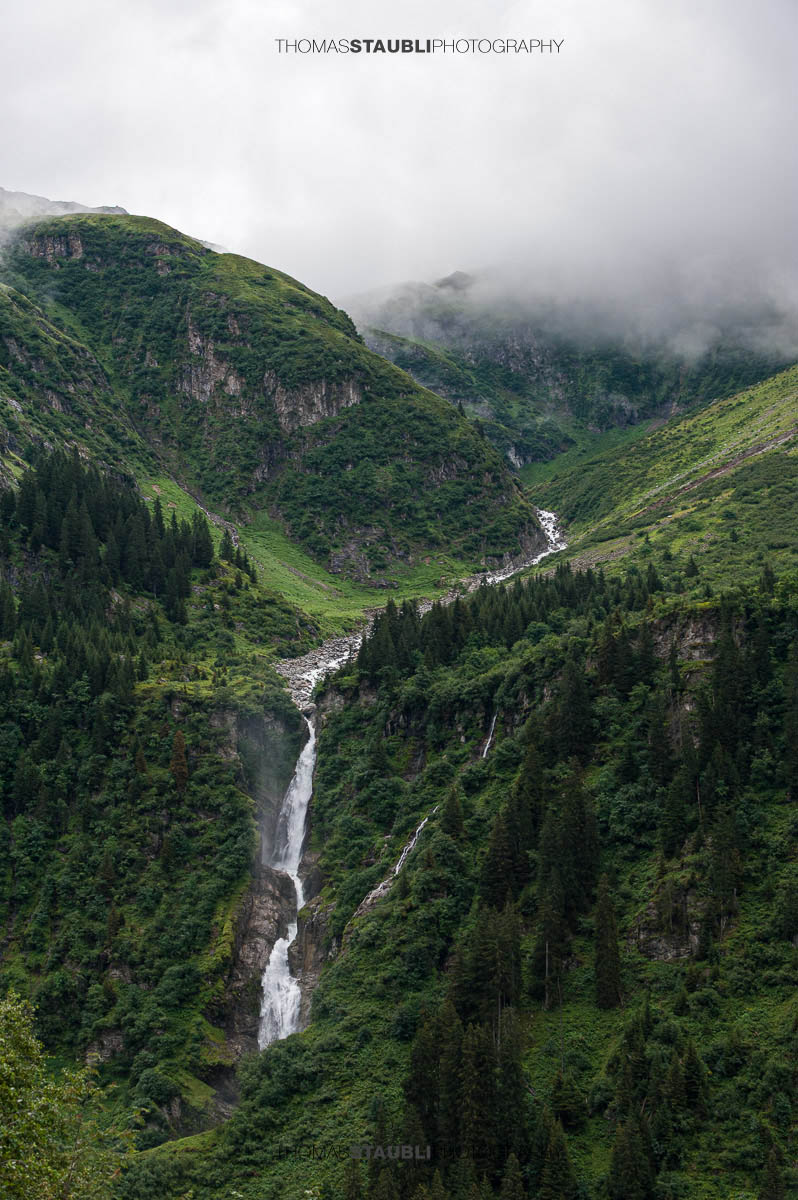 Stäuber Wasserfall im Maderanertal mit tief hängenden Wolken und bewaldeten Berghängen.