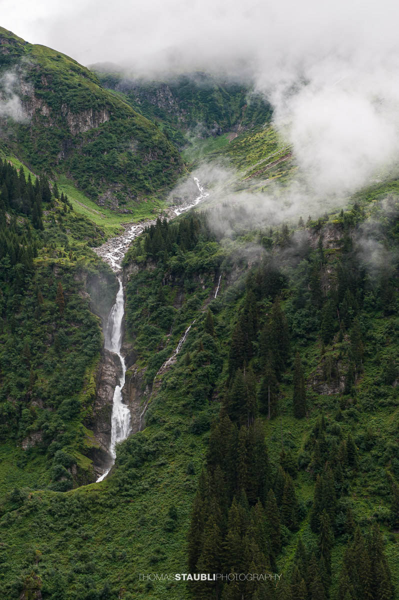 Stäuber Wasserfall im Maderanertal mit tief hängenden Wolken und bewaldeten Berghängen.