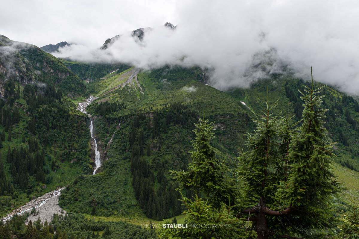 Stäuber Wasserfall im Maderanertal mit tief hängenden Wolken und bewaldeten Berghängen.