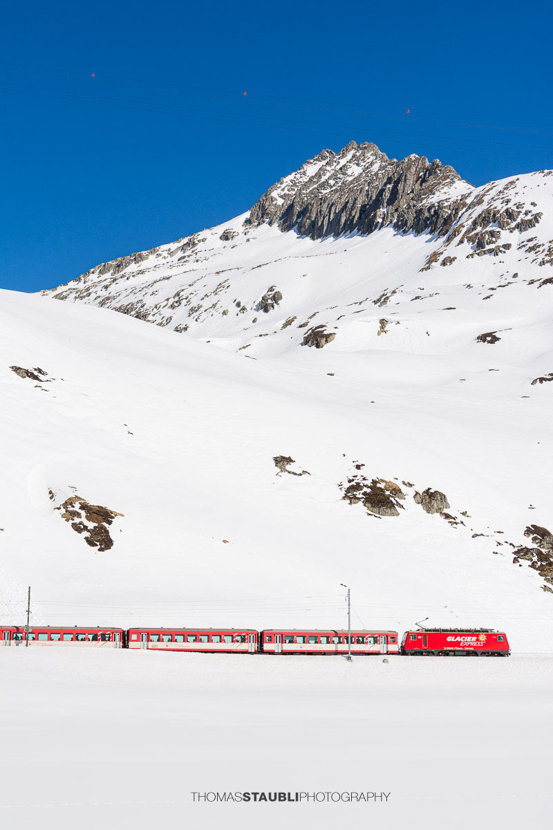 Glacier Express fährt durch die verschneite Landschaft auf dem Oberalppass mit Blick zum Schneehüenderstock.