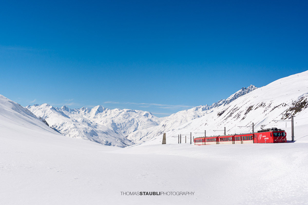 Matterhorn Gotthard Bahn fährt durch die verschneite Landschaft am Oberalppass mit Blick ins Urserental.
